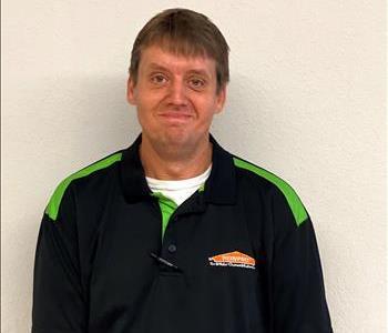 Photo of white male, clean cut brown hair dressed in black and green SERVPRO logo polo shirt against cream colored wall