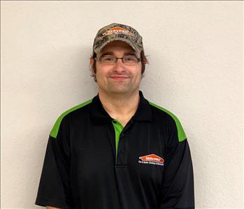 Photo of white male, wearing a logo ball cap, dressed in black and green SERVPRO logo polo shirt against cream colored wall