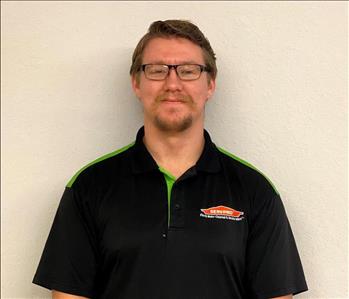 Photo of white male, with glasses, dark hair, dressed in black and green SERVPRO logo polo shirt against cream colored wall